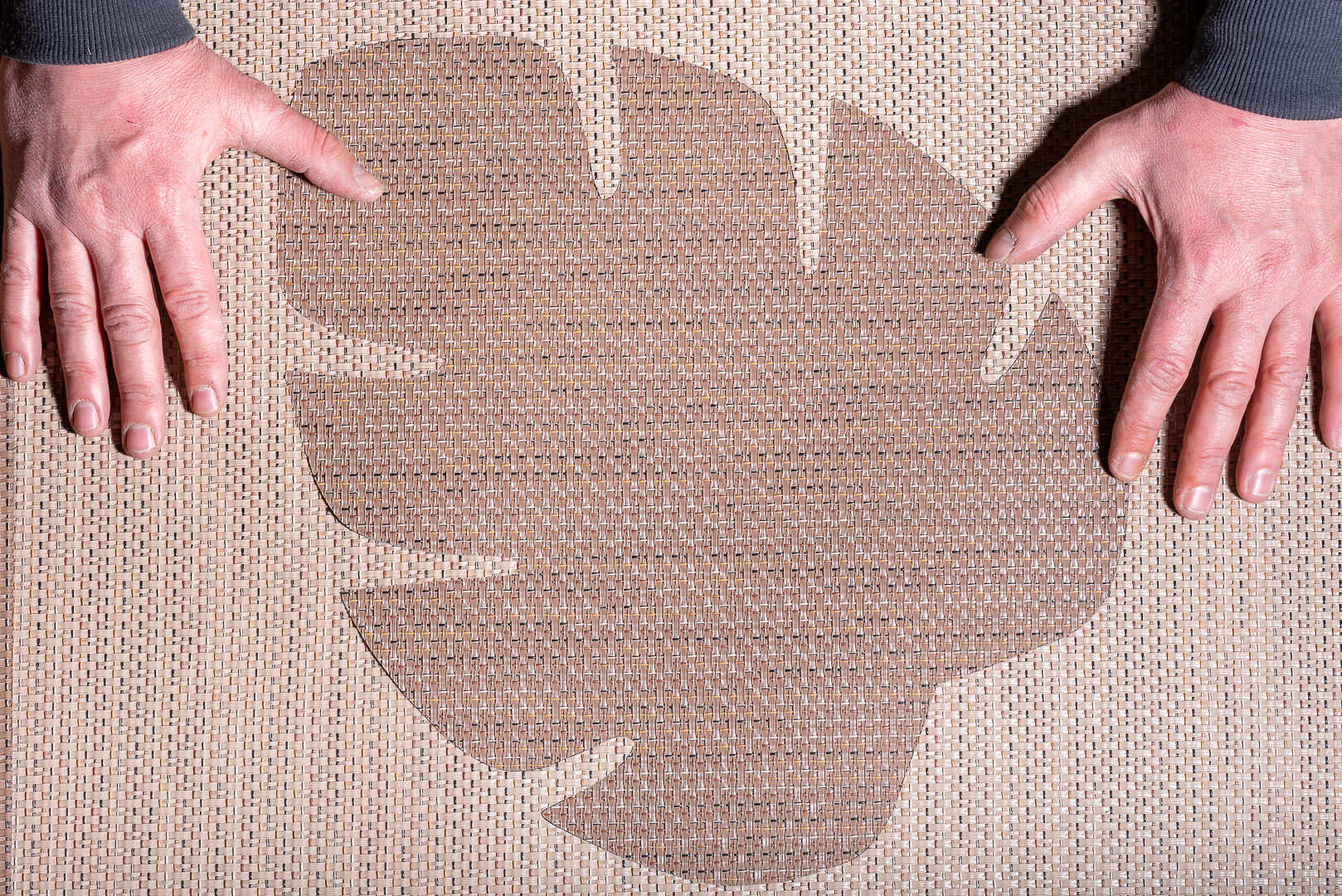 Deux mains sont posées sur un tapis tissé avec une large empreinte en forme de feuille, capturée par l'œil d'un photographe de Villeurbanne. Le tapis présente un motif texturé, et la forme de la feuille se détache sur la surface par ailleurs uniforme, rappelant l'héritage artistique de Lyon.