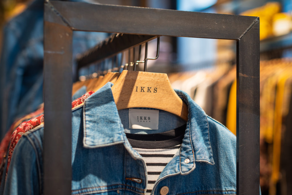 Gros plan sur un portant de vêtements, une veste en jean et une chemise rayée sur des cintres, avec le nom de la marque IKKS. Capturés avec une photographie professionnelle de haute qualité, d'autres vêtements de différentes couleurs sont visibles, flous en arrière-plan, soulignant le style chic de la mode Rhône-Alpes.