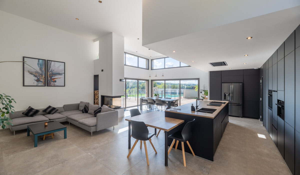 Photographie immobilière d'une cuisine ouverte et un salon modernes et spacieux avec de hauts plafonds et de grandes fenêtres. La pièce comprend un canapé gris, une cuisine noire élégante avec un îlot et une table à manger en bois avec des chaises noires. La lumière pénètre à travers de hautes fenêtres.
