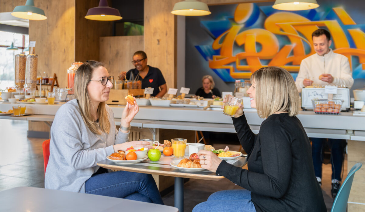 Deux femmes sont assises à une table dans la cafétéria lumineuse et moderne du Photographie Hôtel, savourant leur petit-déjeuner et leurs boissons. En arrière-plan, des clients se servent au buffet, tandis que des graffitis colorés illuminent le mur derrière eux.