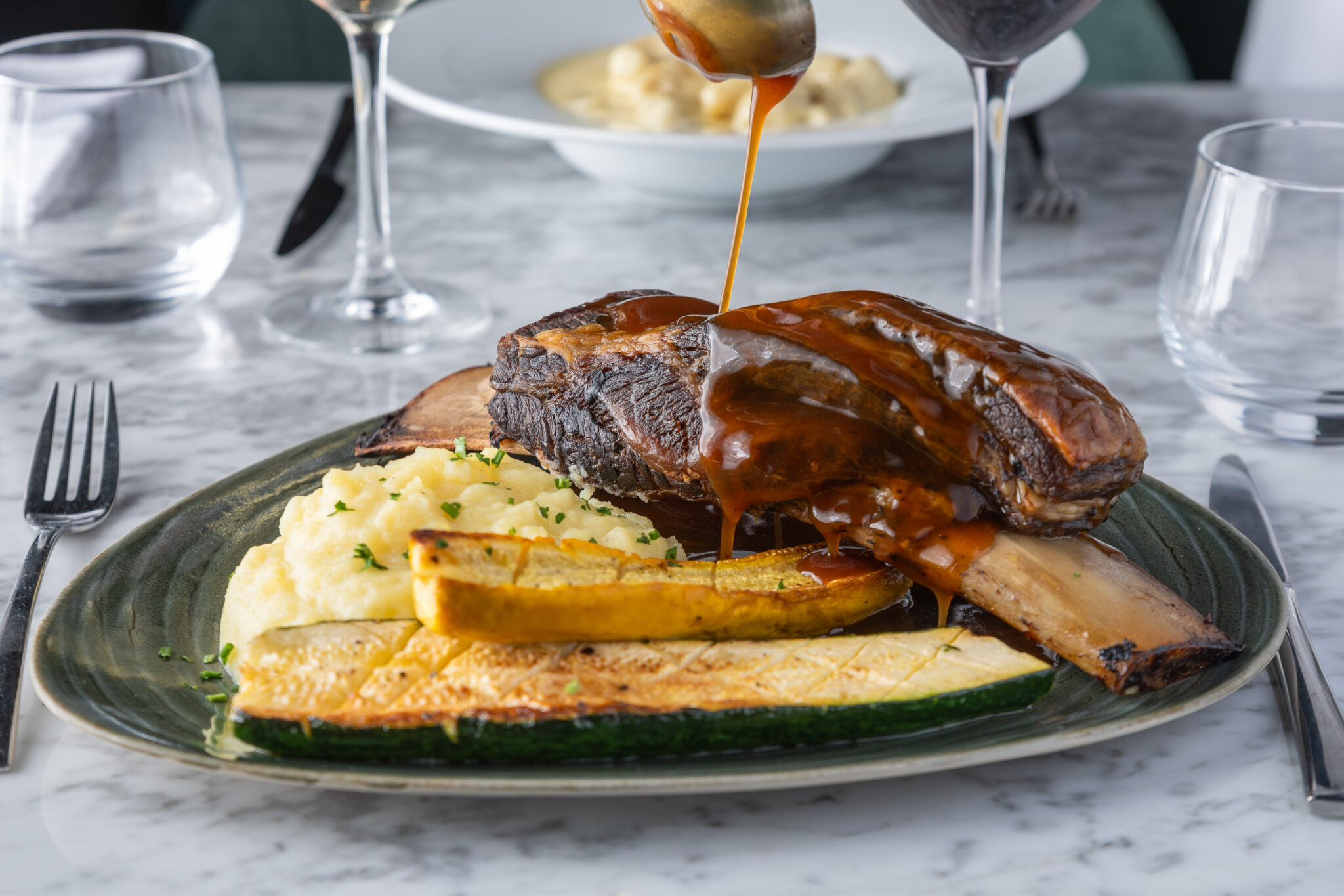 Une assiette de côtes de bœuf braisées nappées de sauce brune, servies avec de la purée de pommes de terre et des courgettes grillées, est astucieusement disposée sur une table en marbre dans le style classique de Photographie Hôtel, avec des verres, des couverts et des pâtes en arrière-plan.