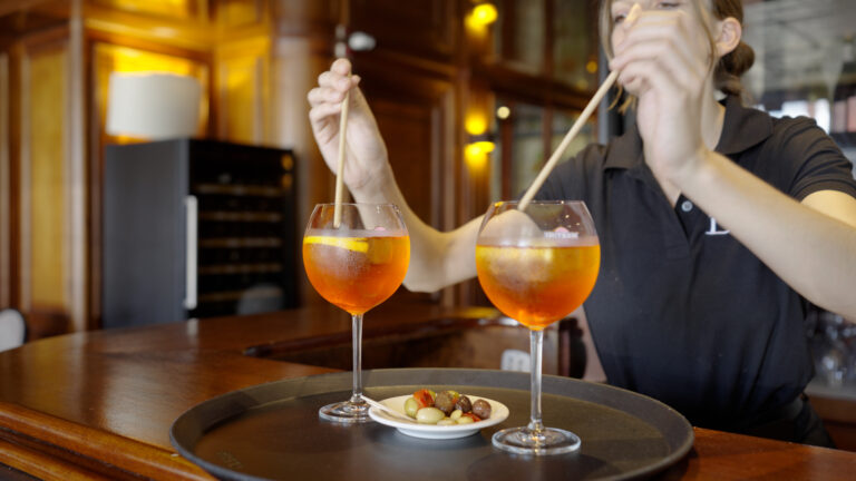 Une personne remue deux cocktails orange dans de grands verres sur un plateau, à côté d'une petite assiette de noix et d'olives mélangées, à l'intérieur d'un bar chaleureusement éclairé avec des intérieurs en bois, photographié par un photographe de reportage basé à Lyon.