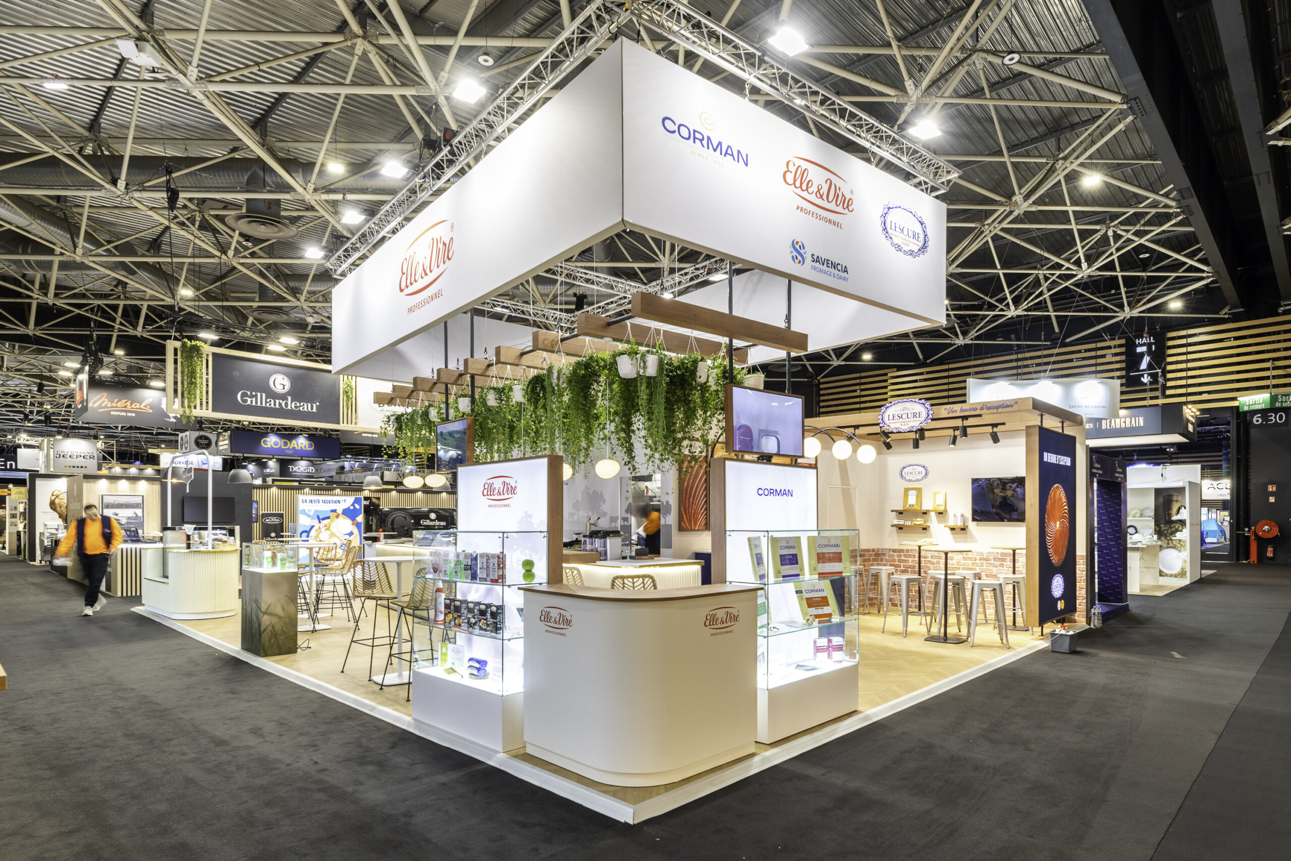 Un stand d'exposition moderne avec un éclairage lumineux, de la verdure suspendue, des vitrines d'exposition et des espaces pour s'asseoir. Le stand présente des marques telles que Corman et Elle & Vire, et est situé dans une grande salle de convention.