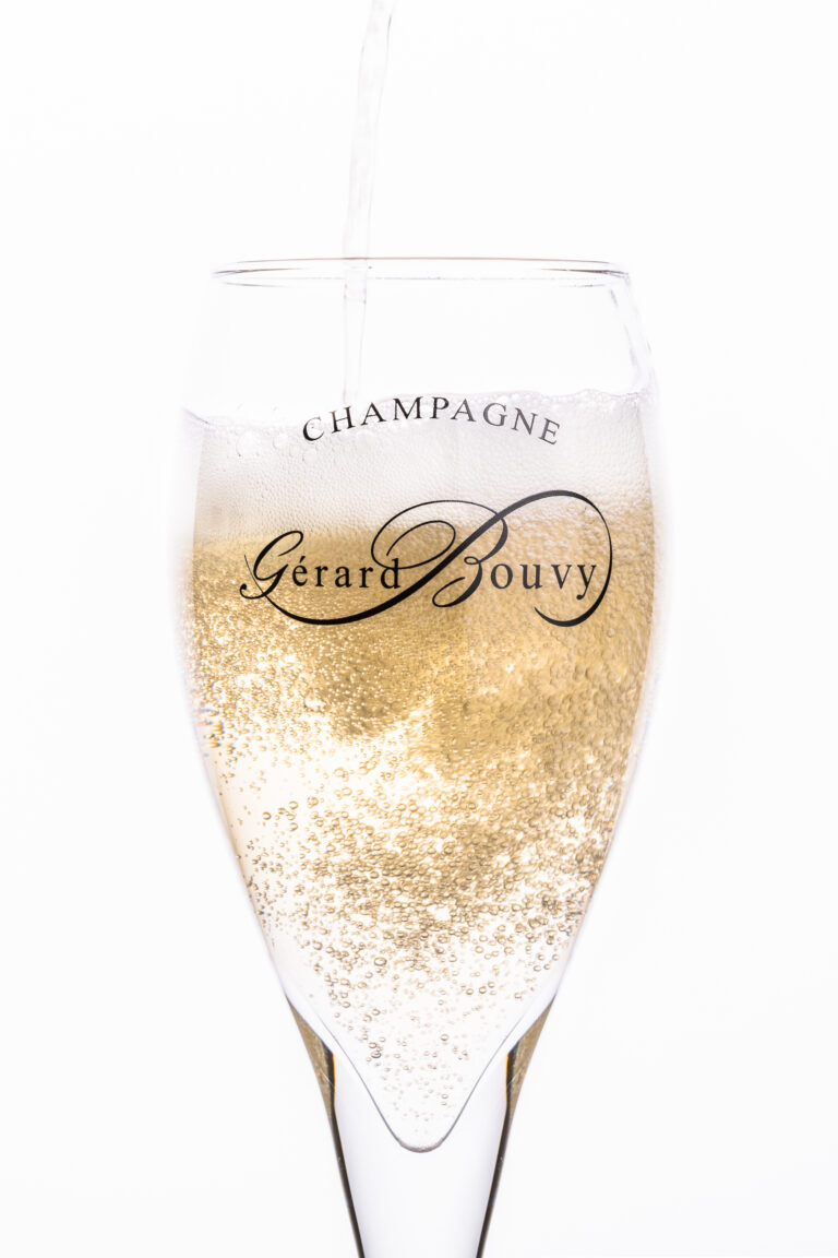Gros plan sur une coupe de champagne étiquetée Champagne Gérard Bouvy, remplie de champagne pétillant, sur fond blanc. Photographe produit Lyon capture les bulles montantes à l'intérieur du verre.