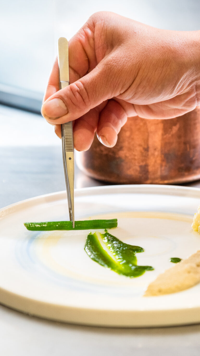 Un gros plan, capturé par un photographe produit Lyon, montre une main munie d'une pince à épiler qui dispose une bande d'aliments verts sur une assiette blanche, créant ainsi un plat minimaliste et artistique rehaussé de purées vertes et beiges.