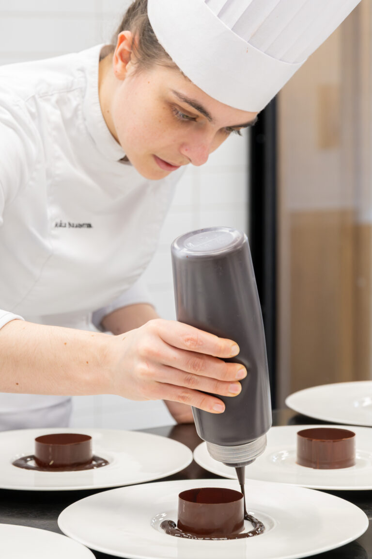 Un pâtissier en uniforme et chapeau blanc presse soigneusement la sauce au chocolat d'une bouteille sur un dessert au chocolat dans une cuisine professionnelle, capturé dans un portrait de reportage par un photographe lyonnais.