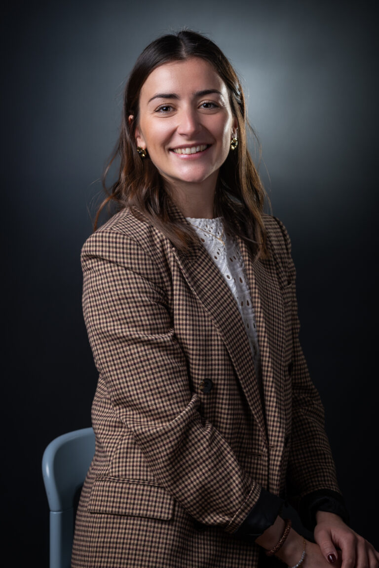 Une femme aux longs cheveux bruns, vêtue d'un blazer à carreaux bruns et d'un chemisier blanc, est assise sur une chaise et sourit à l'appareil photo sur un fond sombre, capturé dans un style rappelant le reportage portrait photographe Lyon.