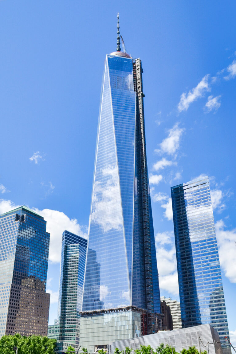 Un grand gratte-ciel moderne en verre, le One World Trade Center, se dresse dans un ciel bleu vif parsemé de nuages. Il est capturé dans un style architectural photographique saisissant et entouré de gratte-ciel dans la ville de New York.