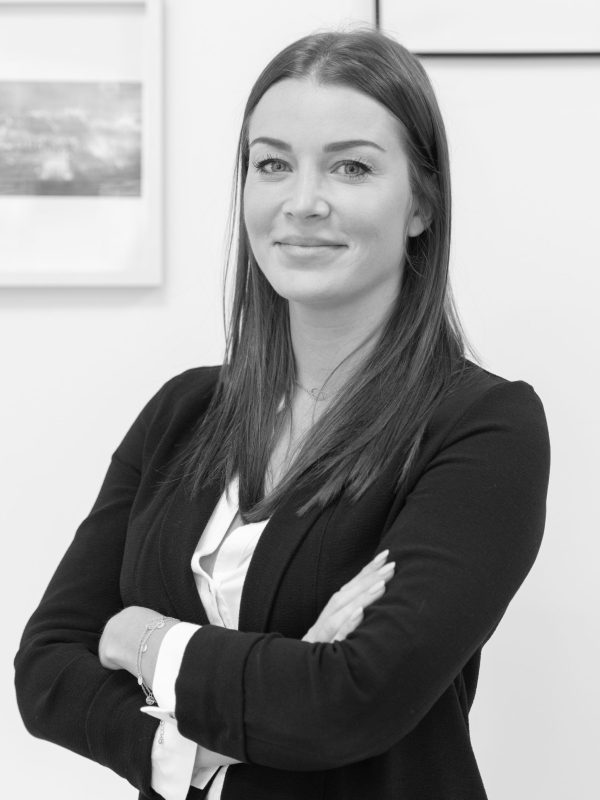 Une femme aux cheveux longs et à l'expression confiante se tient debout, les bras croisés. Elle porte un blazer et un chemisier. L'arrière-plan montre des photos encadrées sur un mur blanc. L'image est en noir et blanc.