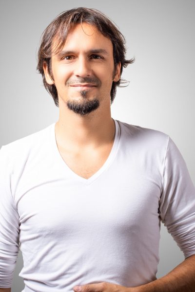 _DSC0160-Edit Un homme aux cheveux mi-longs et au bouc sourit debout sur un fond gris uni. Il porte une chemise blanche à col en V et pose avec sa main posée sur son ventre.