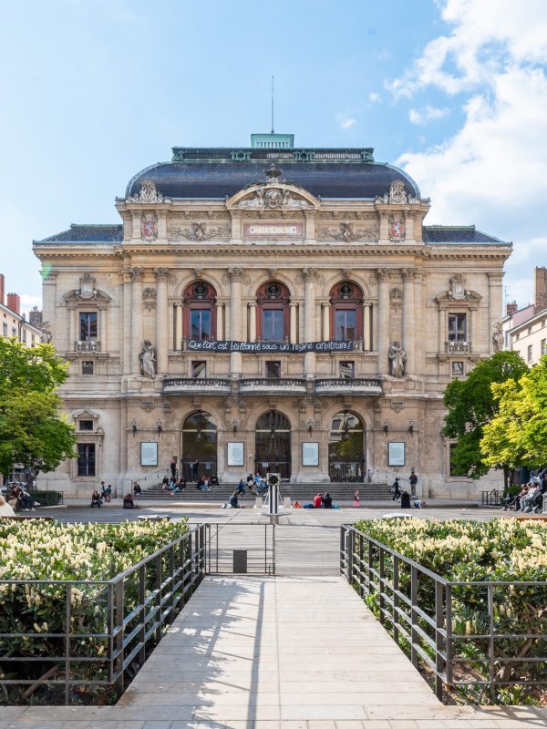 _DSC0313 L'image capture un bâtiment de théâtre historique avec de grands détails architecturaux dans la région Rhône-Alpes, encadré par un chemin bordé d'arbres et de buissons verts luxuriants. Dans cette photographie professionnelle de haute qualité, les gens se promènent et se détendent sous un ciel partiellement nuageux.