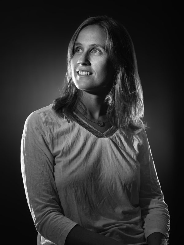Portrait en noir et blanc d'une femme aux cheveux mi-longs, portant un haut clair. Elle regarde vers le haut avec un léger sourire sur un fond sombre avec un éclairage subtil mettant en valeur ses traits.