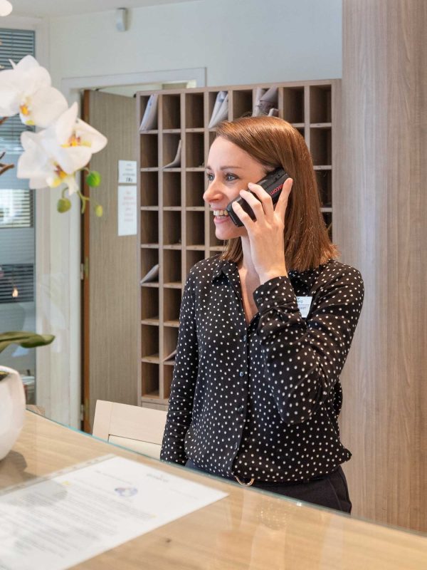 EHPAD_Emera_Villeurbanne_Auvergne-Rhone-Alpes_Personne à l’accueil-1 Une femme portant une chemise noire à pois sourit tout en parlant au téléphone portable. Elle se tient derrière un bureau de réception avec une orchidée en pot à proximité. Derrière elle, il y a un mur de fentes à courrier.