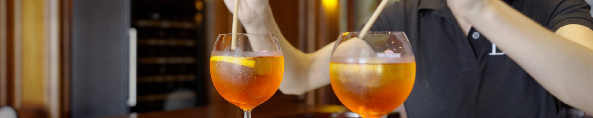 Une personne remue deux cocktails orange dans de grands verres sur un plateau, à côté d'une petite assiette de noix et d'olives mélangées, à l'intérieur d'un bar chaleureusement éclairé avec des intérieurs en bois, photographié par un photographe de reportage basé à Lyon.