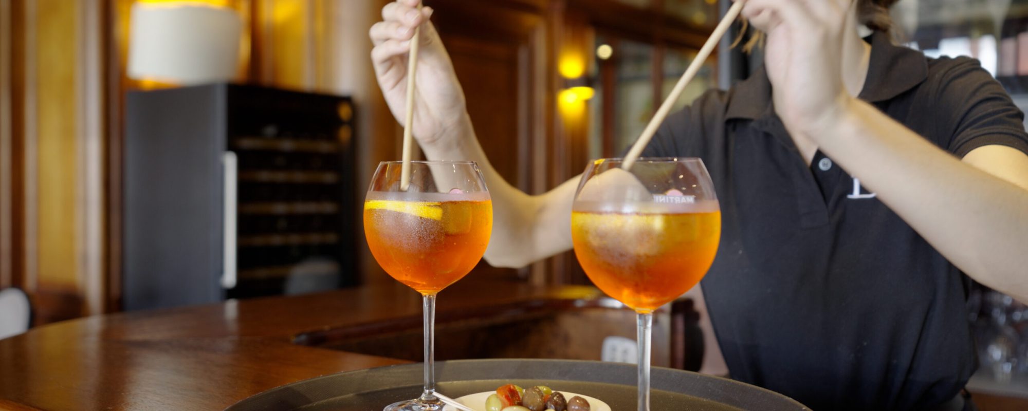 Une personne remue deux cocktails orange dans de grands verres sur un plateau, à côté d'une petite assiette de noix et d'olives mélangées, à l'intérieur d'un bar chaleureusement éclairé avec des intérieurs en bois, photographié par un photographe de reportage basé à Lyon.