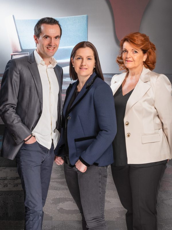 Trois personnes posent pour un portrait, capturé par un photographe expérimenté à Villeurbanne. L'homme à gauche porte un costume gris, la femme au centre arbore un blazer bleu et la femme à droite porte une veste blanche. Derrière eux, des formes abstraites dansent dans une harmonie doucement éclairée.